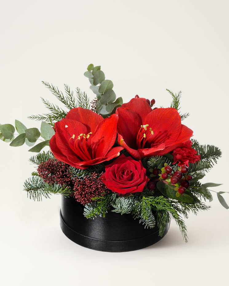 Önska God Jul med ett blommogram från Interflora! Rund svart ask, med ett blomsterarrangemang bestående av röda snittamaryllisar, röda rosor och dekorationsgrönt.