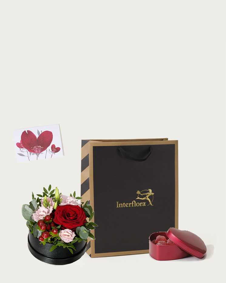 Presentpåse med blomsterdekoration i rund liten hattask (röda rosor, prärieklocka, hypericum, nejlikor och dekorationsgrönt). Plus en ask med röda geléhjärtan och ett fint gratulationskort.