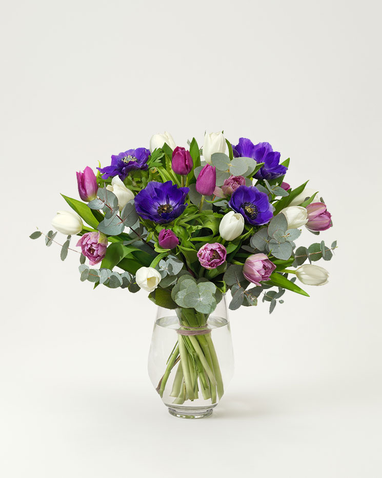 Ståtlig vårbukett med blommor i blått, vitt och lila tillsammans med gröna blad( tulpaner, anemoner och eukalyptus). Buketten finns att beställa online hos Interflora.